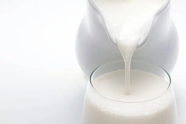 Milk being poured from a white ceramic pitcher into a glass on a white background.
