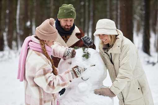 Happy family of father, mother and daughter making snowman while standing in front of camera during stroll in winter park or forest