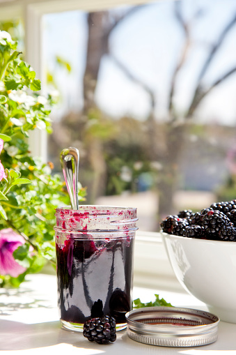 Black raspberry jam in a glass jar by a window.