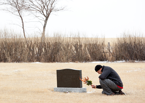 A man pays his respects at a grave. A man dressed in black for a funeral is bowing or praying at a gravestone. Themes include dying, death, grief, loss, grieving, sadness, tears, remembering, veterans day, memorial day, and lest we forget. Horizontal colour image taken in mid winter. Man is in his 30s, side view, Caucasian. 