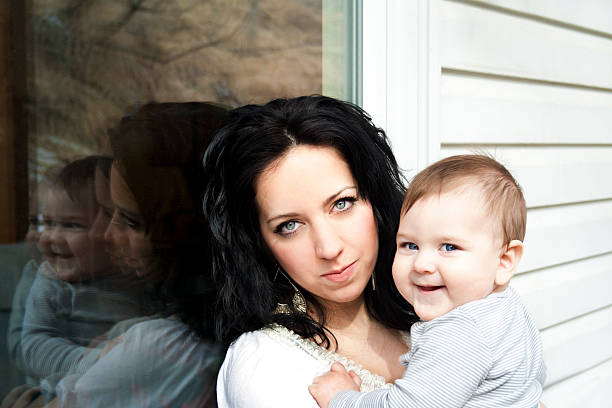 Mother holds baby while leaning against window stock photo