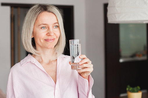 Close up of thirsty adult Caucasian female hold glass drink clean mineral water for body refreshment and healthy skin. Middle aged 40s woman enjoy clear pure aqua feeling dehydrated. Hydration concept