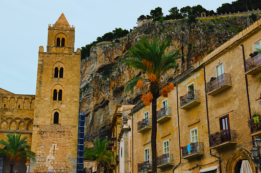 Landscape view of old city in Cefalu. Tower of ancient Cathedral of Cefalu and old residential house. Rocky mountain with green trees in the background. Travel and tourism concept. Cefalu, Sicily.