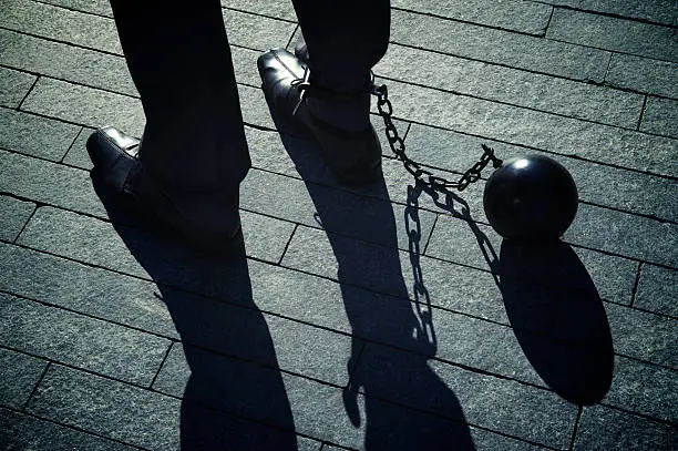 "Long, dark shadows hang from a the feet of a businessman standing with iron ball and chain"