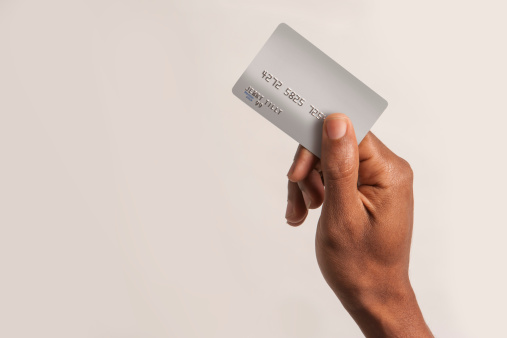 An African American  man's hand holding a fake credit card with fake numbers and a fake name on it. Photographed against an off white background.