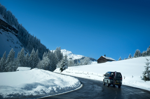 Mini van car with ski's mounted on a rear rack driving through snow covered alpine valley with wooden chalets towards ski slopes and mountains.