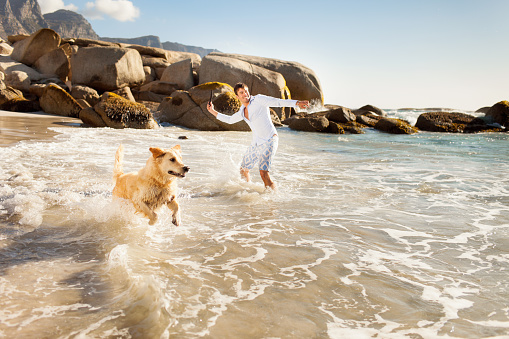 A horizontal shot of a happy young man playing with his dog on a beach.  The man has dark hair, and he is dressed in white.  There is a foamy tide in the foreground and a configuration of rocks and blue sky in the background.