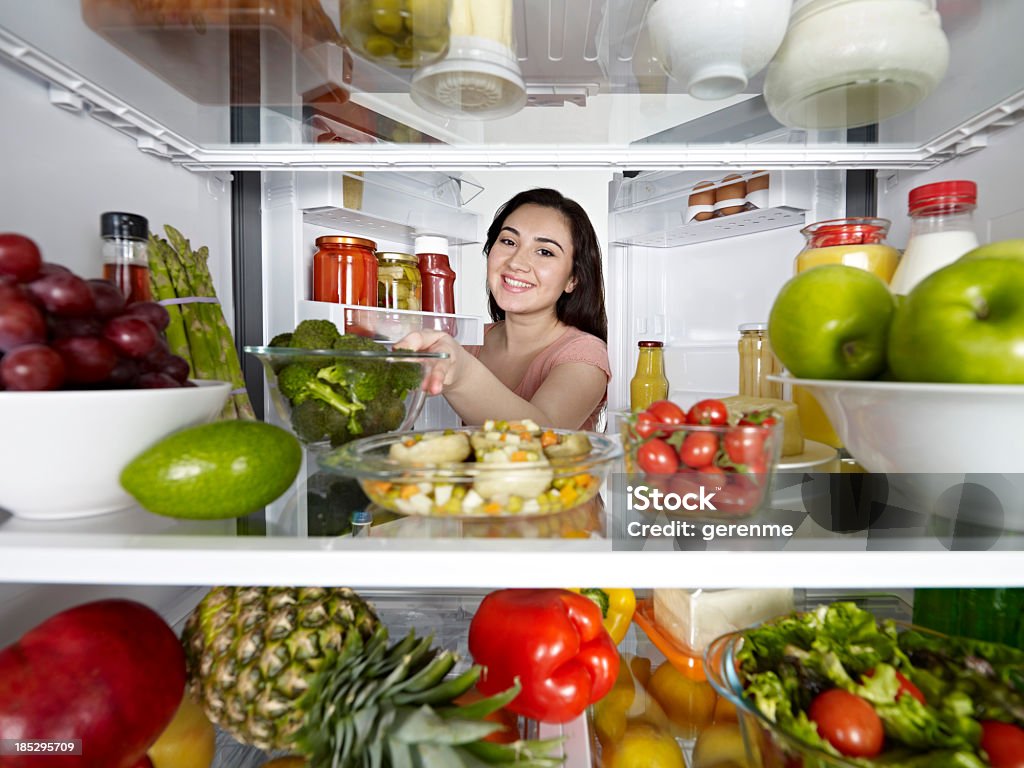 Taking healthy food from fridge Looking from inside a fridge full of healthy food to a young woman smiling to the camera Refrigerator Stock Photo