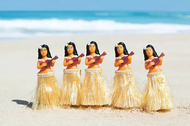 A group of five Hawaiian hula dancer figurines doing the traditional hula dance performance and playing the ukulele on a Hawaiian Island beach on the island of Kauai, Hawaii, USA. Pacific ocean waves are in soft focus in the background. Photographed in a horizontal format with copy space.