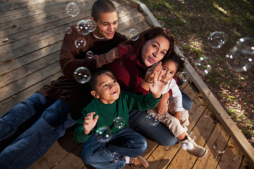 Portrait of Hispanic family at a park playing with bubbles.  Parents 20s, children 2 and 5 years.