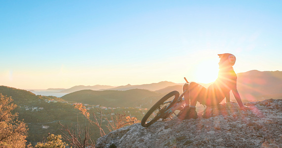 Mountain biker relaxes on mountain outcrop above hills and sea at sunset