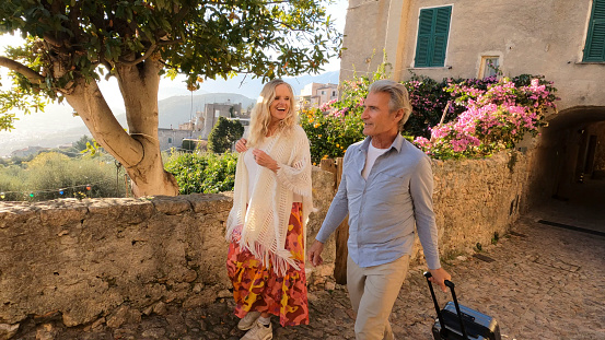 Upscale couple pull luggage through old village, lush gardens and hills distant