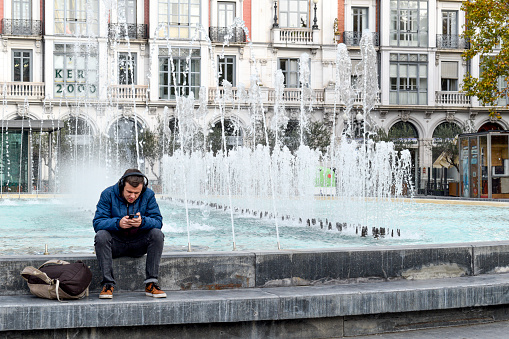 Valladolid- Spain , December 2023 ,
A young man is sitting in a fountain, in the center of the city, listening to music with headphones, using the cell phone - Valladolid - Spain