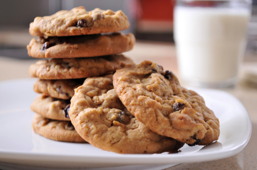 Horizontal image of a stack of warm oatmeal raisin cookies sitting on a white plate on a kitchen counter. A glass of cold milk sits in the background. Focus in on the center of the 2 cookies in the foreground.