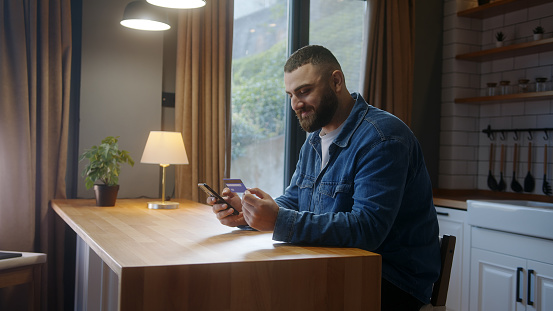 Bearded young adult man sitting against the kitchen counter entering credit card number on smartphone for makes secure easy distant electronic payment at home
