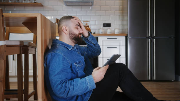 istock Young man sitting on the floor leaning against the kitchen using tablet device having anxiety and stress. Upset man reacting to loss, bad news 1853069179