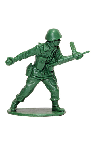 A green toy soldier against a white background. This soldier is cradling a machine gun while lobbing a grenade.