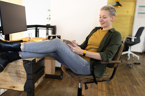 Relaxed woman using on mobile phone at work with feet up on desk. Young businessman sitting in office with feet on table using smart phone during break.