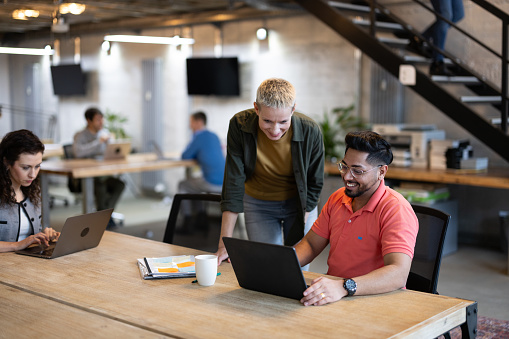 Business people working at open plan startup office. Businesswoman standing by male colleague working on laptop at coworking office.