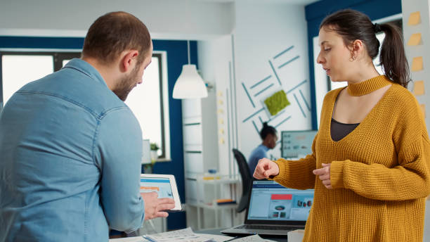 istock Man in startup office zoomin in digital tablet using touchscreen presenting sales charts to woman colleague 1853145857