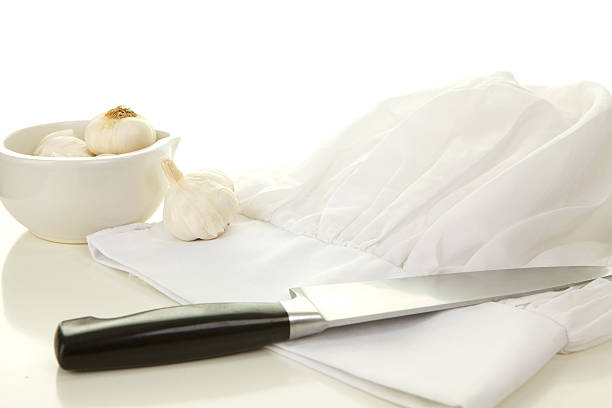 A cooking background with a flat white chef hat on a white table with a kitchen knife  and a  bowl with garlic bulbs.  Shot in the studio in front of a white background