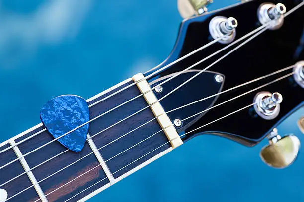 Blue guitar pick between strings over rosewood fretboard and against nature blue background.