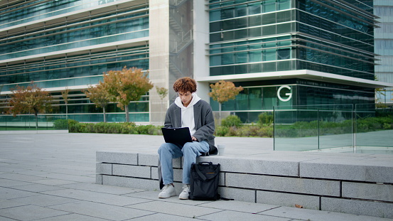 Focused freelancer typing laptop on street. University student learning online