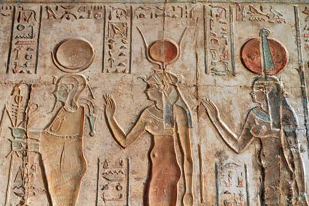 "Painted sunk relief from the Ptolemaic Temple of Hathor at Deir el-Medina, part of the Theban Necropolis, Luxor, Egypt.  The relief depicts three gods.  On the left is Osiris, who wears the lunar disk.  Hathor, goddess of love music and dance is in the middle and wears the solar disk and cow horns.  The figure on the right may be Isis.  Although she is not wearing the throne-shaped headgear with which she is normally associated, the feather suggests that it is."