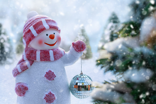 Happy snowman decorates Christmas tree with Christmas bauble on a snowy winter day. Winter holidays background. Greeting card. Fabulous Christmas composition with snowman dressed in warm clothes, mittens, scarf and hat.