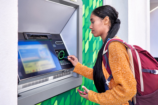 Young and pretty digital nomad Colombian Latin girl with her backpack scanning direro with her credit card at an ATM
