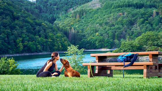 Young woman resting with her dog on green meadow by blue lake and green mountains. Travelling with dog