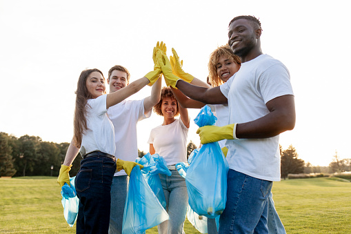 group of multiracial people in gloves with garbage bags high-fiving and celebrating success together in the park, team of volunteers cleaning up trash and rejoicing in achievements