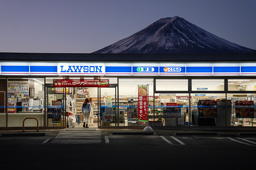 Kawaguchiko, Japan - December 7, 2023: Customers entering a Lawson konbini (convenience store) with Mount Fuji seen behind it at dawn.