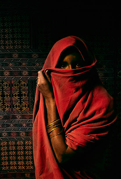 A north African woman covers her face with a veil. She is wearing wrist bangles and standing in front of a kilim rug.   Scanned from film.