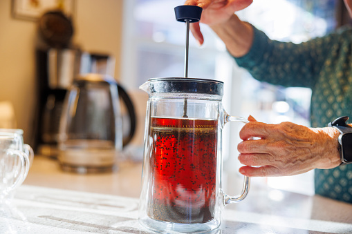 Unrecognizable senior woman hands using French Press to steep tea on bright kitchen counter