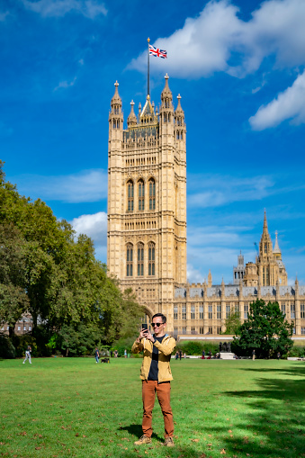 Young Latin man in casual clothes smiling and using smartphone on background of Big Ben against clock tower in London