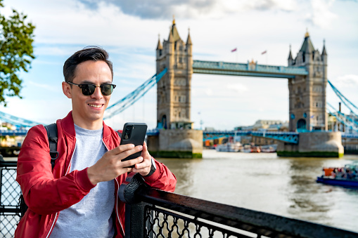 Young Latin man wearing sunglasses using smartphone near Tower Bridge in London