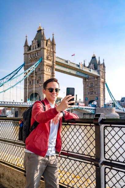 istock Happy young Latin man wearing sunglasses and red jacket with backpack taking selfie with smartphone near metal railing of famous Tower Bridge in London 1853689914