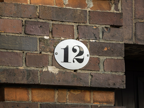 Number 12 on an exterior house wall. The twelve is part of an address. The metal plate is white with black figures. The design is vintage and old fashioned.