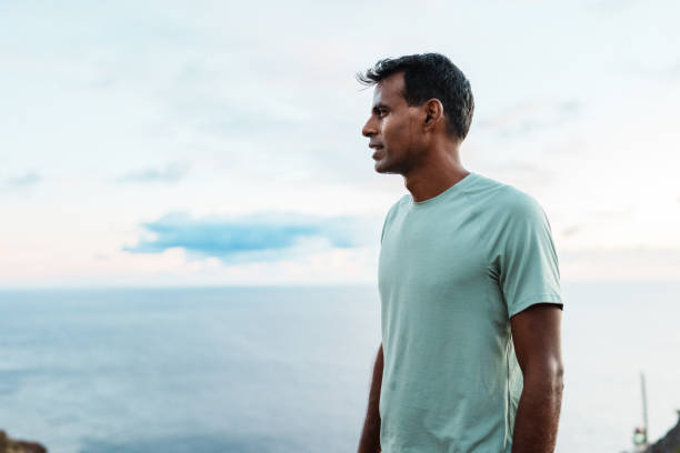 An active and healthy man of Indian descent who is hiking solo along the coast of Hawaii at sunrise pauses along the trail to take in the scenic ocean views.