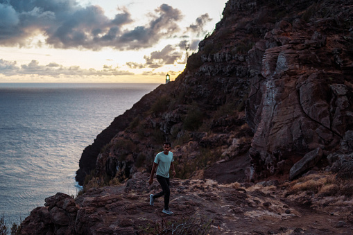 An active and healthy man of Indian descent hikes along a rocky trail along the coast of Hawaii overlooking the Pacific Ocean at sunrise.