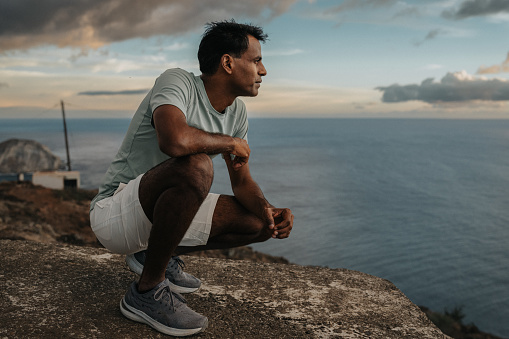 An active and healthy man of Indian descent who is hiking solo along the coast of Hawaii at sunrise pauses along the trail to take in the scenic ocean views.