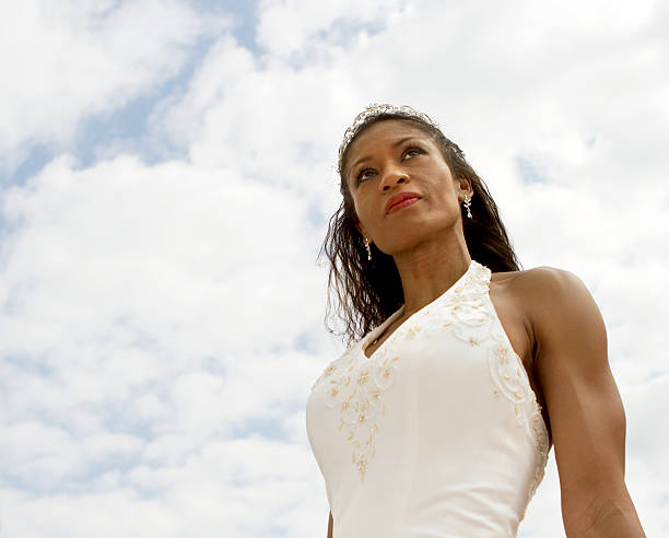 A bride is getting ready for her wedding and is wearing a white dress. The dress is flowing and has a veil on it. The bride is standing in front of a bed, and the bed is covered with a white sheet