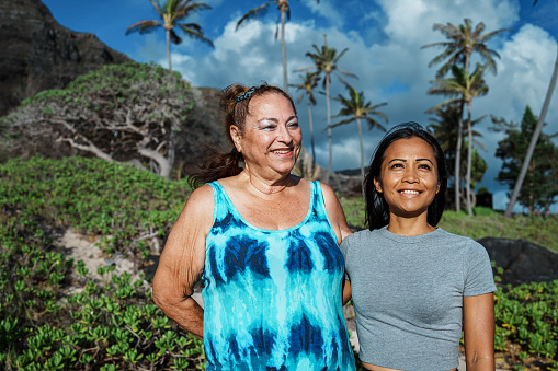 Portrait of a vibrant and joyful multiracial senior woman and her beautiful adult daughter smiling and embracing while standing on the beach in Hawaii and looking out at the ocean.