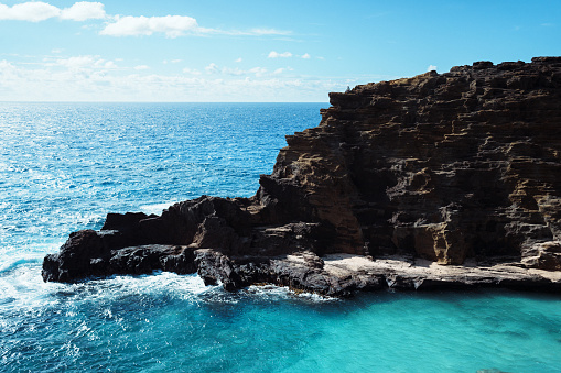 Wide shot of an active man of Indian descent sitting atop a rocky cliff with a scenic view of aquamarine ocean water off the coast of Oahu in Hawaii.