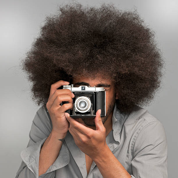 head and shoulders shot of a man with big afro hair holding a vintage camera taking a picture on gray background