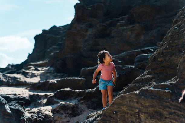 An elementary school age girl of multiracial ethnicity and her siblings hike and climb a rock structure while spending time adventuring outside.