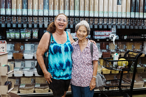 A senior woman of Korean descent and a multiracial senior adult friend who are shopping at the grocery store together embrace and smile toward the camera.