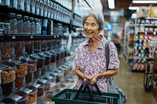 An elderly woman of Korean descent holds a shopping basket and smiles directly at the camera while shopping in the bulk foods section of an organic health foods grocery store in Hawaii.