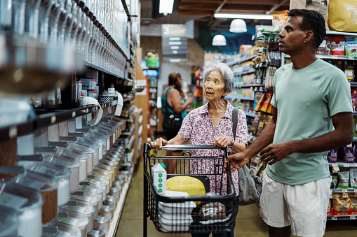 An elderly Korean woman shops in the bulk foods section at an organic grocery store with the help of a volunteer caregiver of Indian descent. The care provider is working for a community outreach program aimed to provide support and nutrition guidance to senior adults.